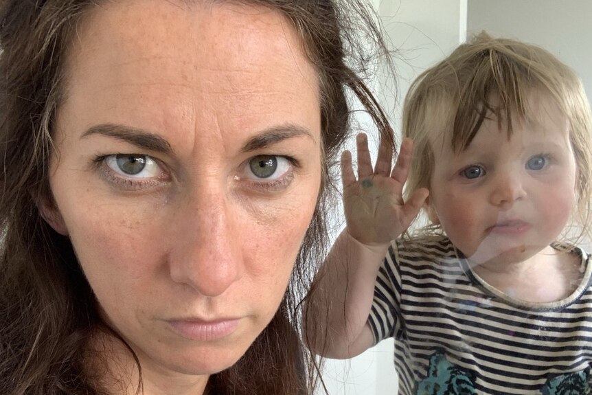 Woman stares blankly at the camera. Next to her a toddler squishes her face against a glass window