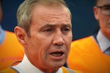 A man speaking with some people wearing orange high vis vests in the background.
