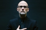 bald man with short beard wears a dark suit and glasses. he holds a hand over his chest.