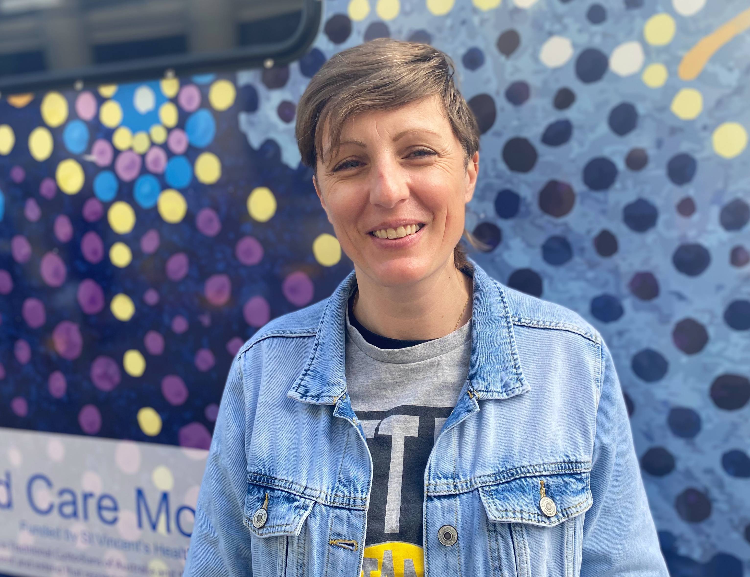 A young woman with short dark blonde hair, wearing two T-shirts and a denim jacket smiles as she looks directly at the camera