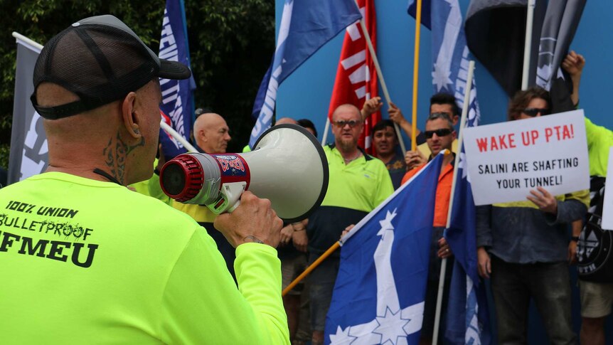 A worker in a high-viz green shirt holds a megaphone in front of a group of protesters holding flags and signs.