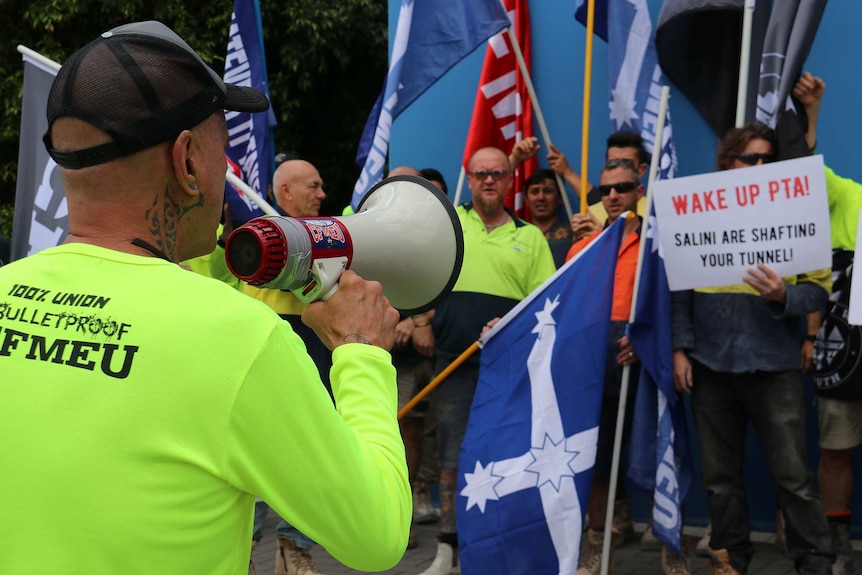 A worker in a high-viz green shirt holds a megaphone in front of a group of protesters holding flags and signs.