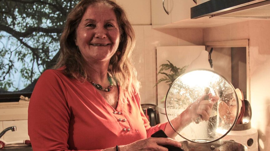 A woman holds a saucepan lid over her stove while a bright light shines through it. She's smiling for the camera.