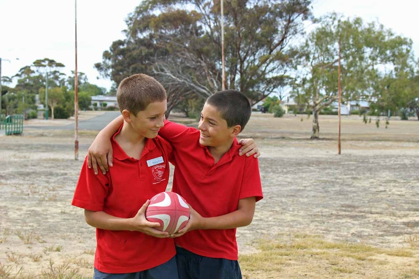 Two boys play with a football in front of old goalposts on a dirt oval.