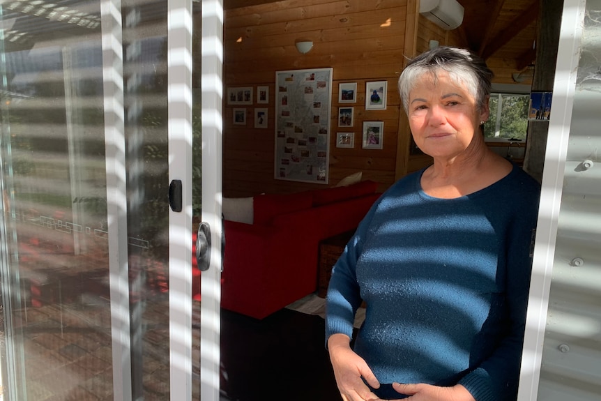 A middle-aged white woman with short grey hair. She is standing by a door, wearing a blue sweater