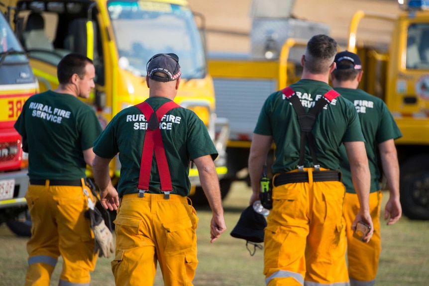 Four firefighters with 'Rural Fire Service' written on the back of their shirts walk away from the camera towards fire trucks.