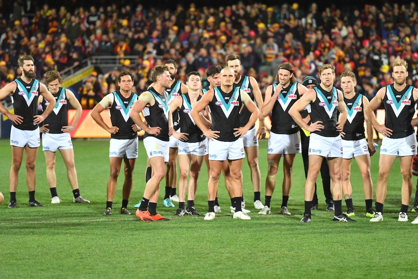 Port Adelaide players stand in a line looking upset with Adelaide fans in the background