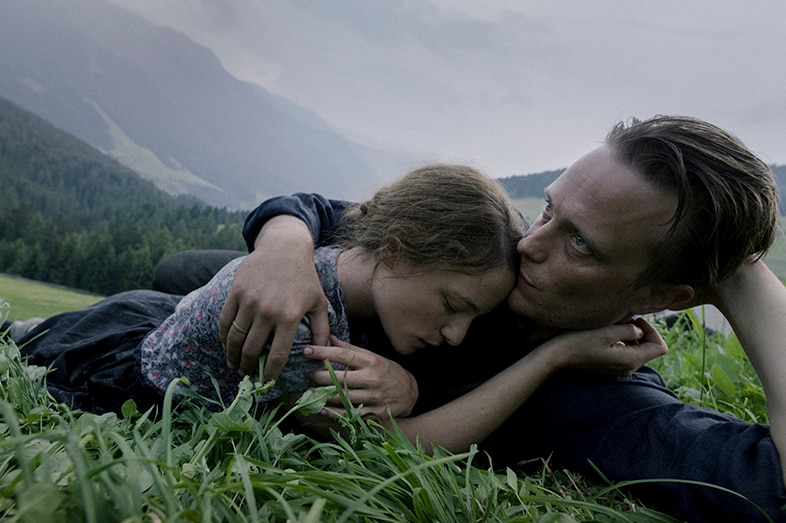 A woman with eyes closed and man with serious expression lay in an embrace on grass in the countryside on an overcast day.