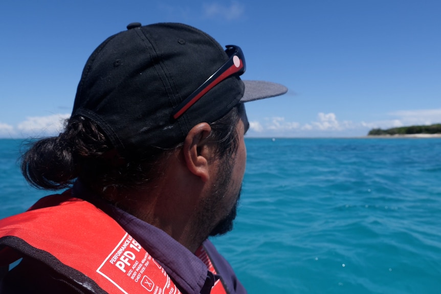 Side profile of Jacob with a cap on, Aboriginal flag sunglasses on his head, hair tied back, looking towards island, blue sea.