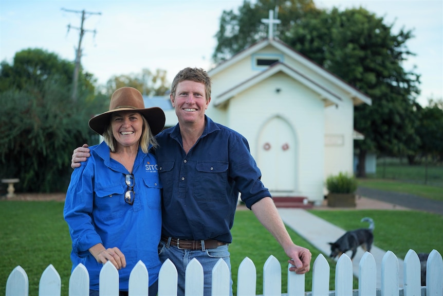 Woman in a blue button-up shirt and wide brim hat stands with a man in navy button up shirt in front of a small white chapel 