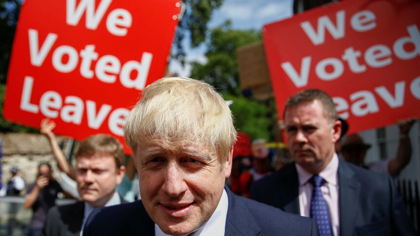 A blond man in a suit smiles in front of posters reading "We Voted Leave"