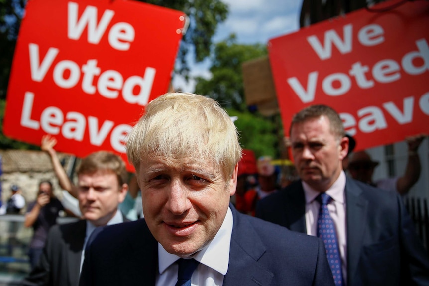 A blond man in a suit smiles in front of posters reading "We Voted Leave"