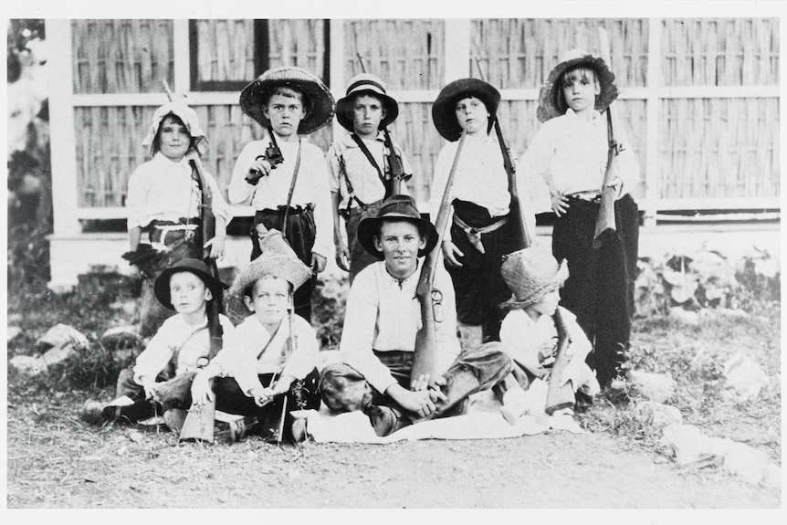 A black and white photo of nine children sitting on a dirt lawn bearing rifles.