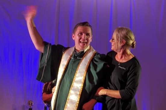 Tristan Sik wearing acamedic robes stands with a support worker and lifts his arm in celebration 