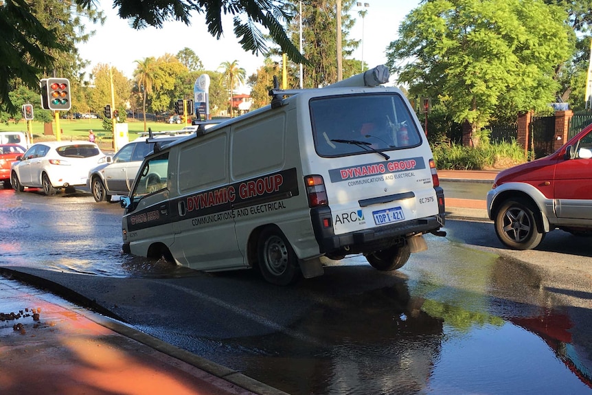 The front wheels of a van are stuck in a deep puddle after a water main bursts.