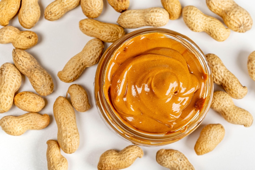 A glass jar of peanut butter surrounded by peanuts