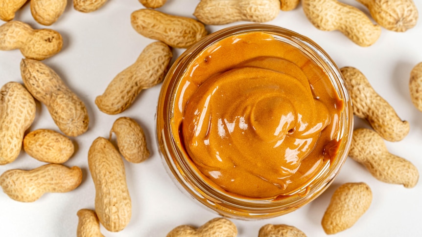 A glass jar of peanut butter surrounded by peanuts