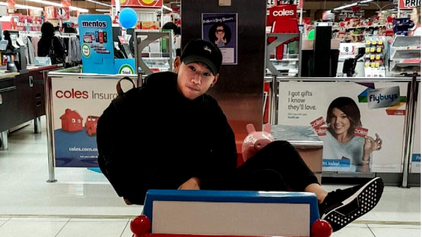 Dillon Wu, wearing a black hoodie, pants and cap, sits in a small toy car in front of a supermarket.