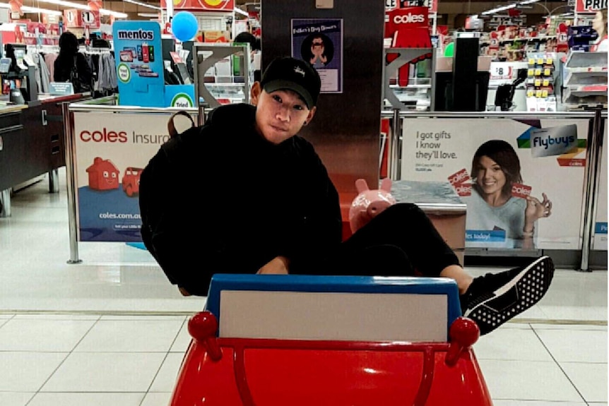 Dillon Wu, wearing a black hoodie, pants and cap, sits in a small toy car in front of a supermarket.