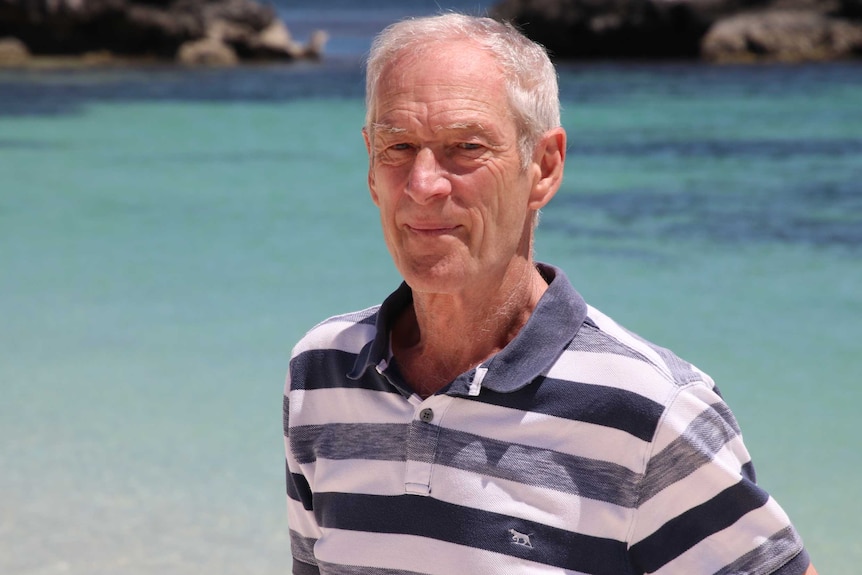 Ian MacRae stands on a beach with turquoise blue water in the background.