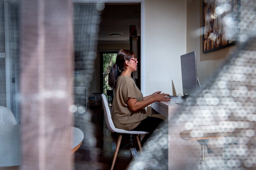 Sandy Trujillo sits at her desk, looking at a computer. She is seen through a fly wire door.