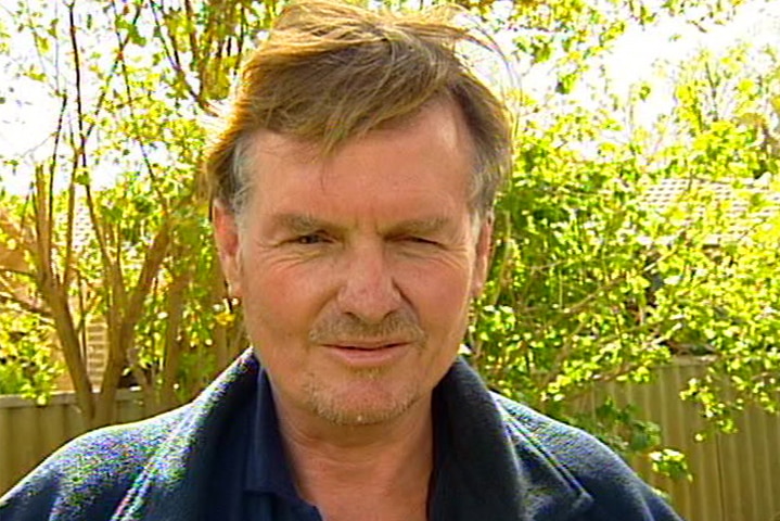 Head and shoulders shot of an unshaven, middle aged man standing in a yard with trees behind him.