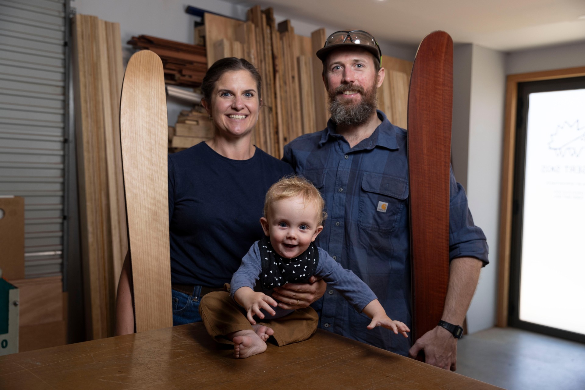 A man and a woman holding skis in a workshop setting, with a small baby.