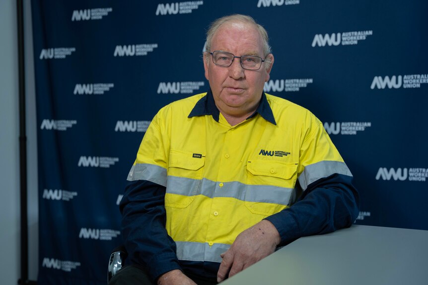 A man sits in front of a navy banner in a high vis shirt, looking at the camera.