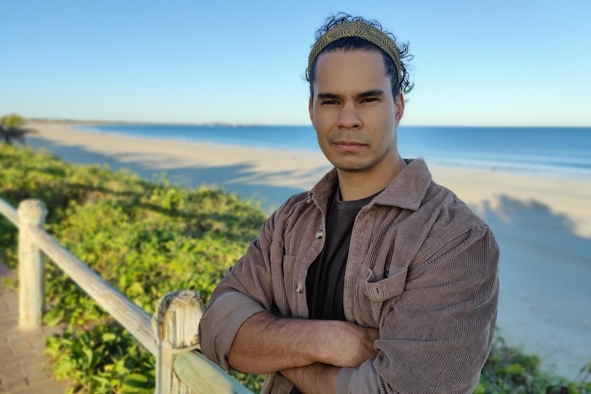 An Aboriginal man with a bandana stands with his arms folded in front of a beach
