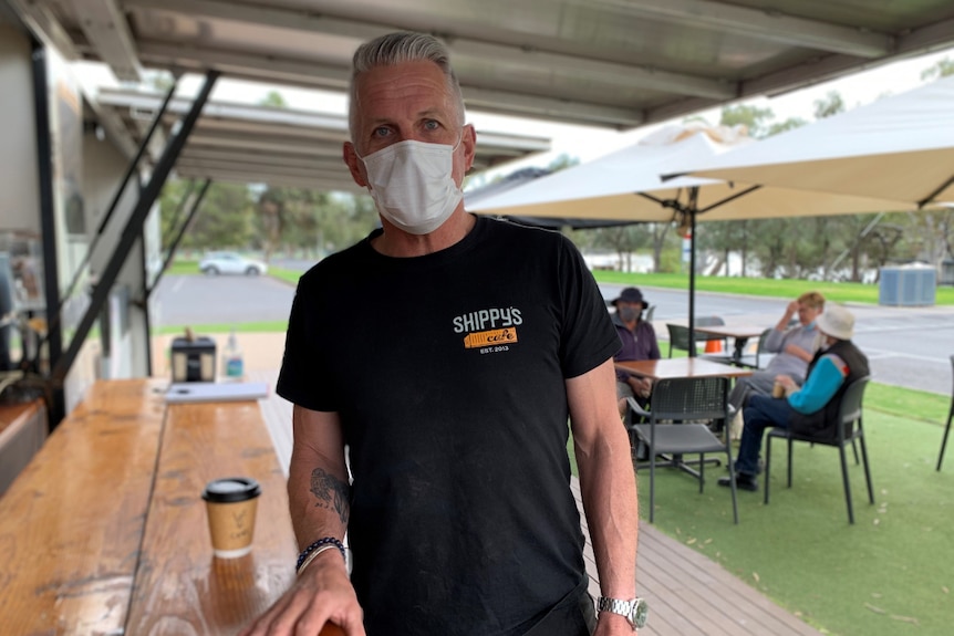 A man looks at the camera with a mask on. Customers sitting down at a table in the background.