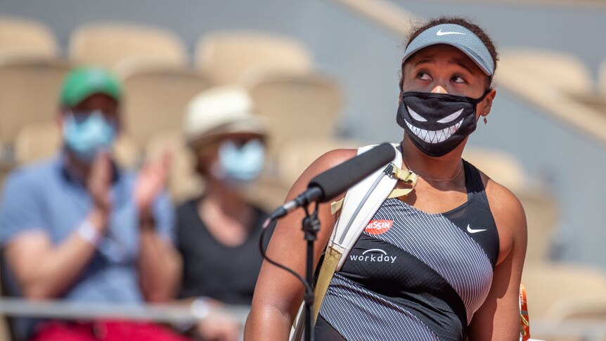 Tennis player Naomi Osaka standing on a tennis court with a microphone in front of her. She is wearing a mask.
