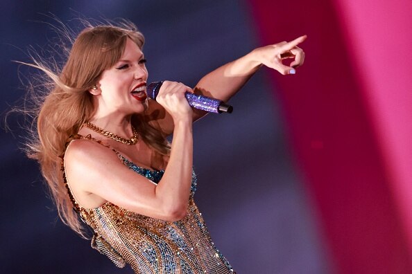 Taylor Swift, a singer with long blonde hair holds a microphone in one hand and points with the other