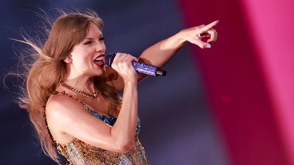 Taylor Swift, a singer with long blonde hair holds a microphone in one hand and points with the other