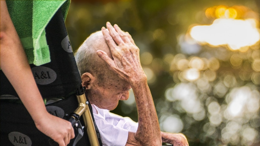 An old man in a wheelchair holds his head.