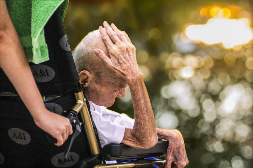 An elderly man in a wheelchair with his hand on his head, with a carer.