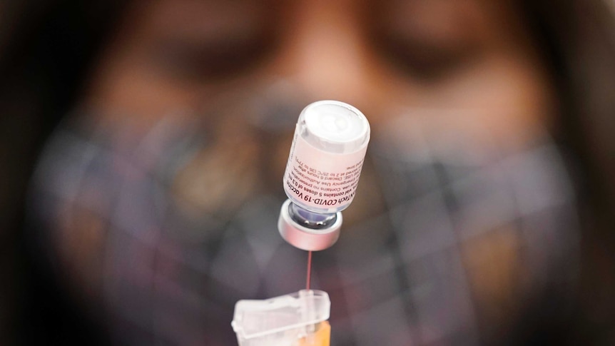 A vial of Pfizer BioNTech vaccine is seen upside down and suspended at the end of a syringe, with a nurse's face behind it