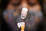 A vial of Pfizer BioNTech vaccine is seen upside down and suspended at the end of a syringe, with a nurse's face behind it