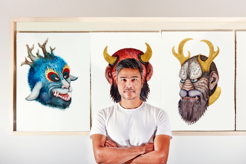 Artist Khadim Ali standing in front of paintings of three fantastical characters, his head framed by devil horns