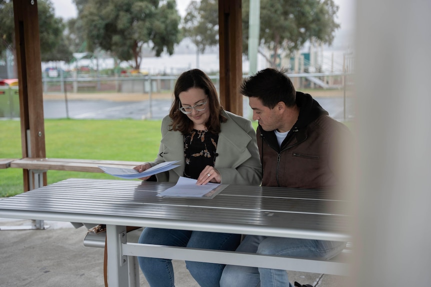 A man and woman sit at a metal picnic table in a park under a pergola reviewing A4 documents.