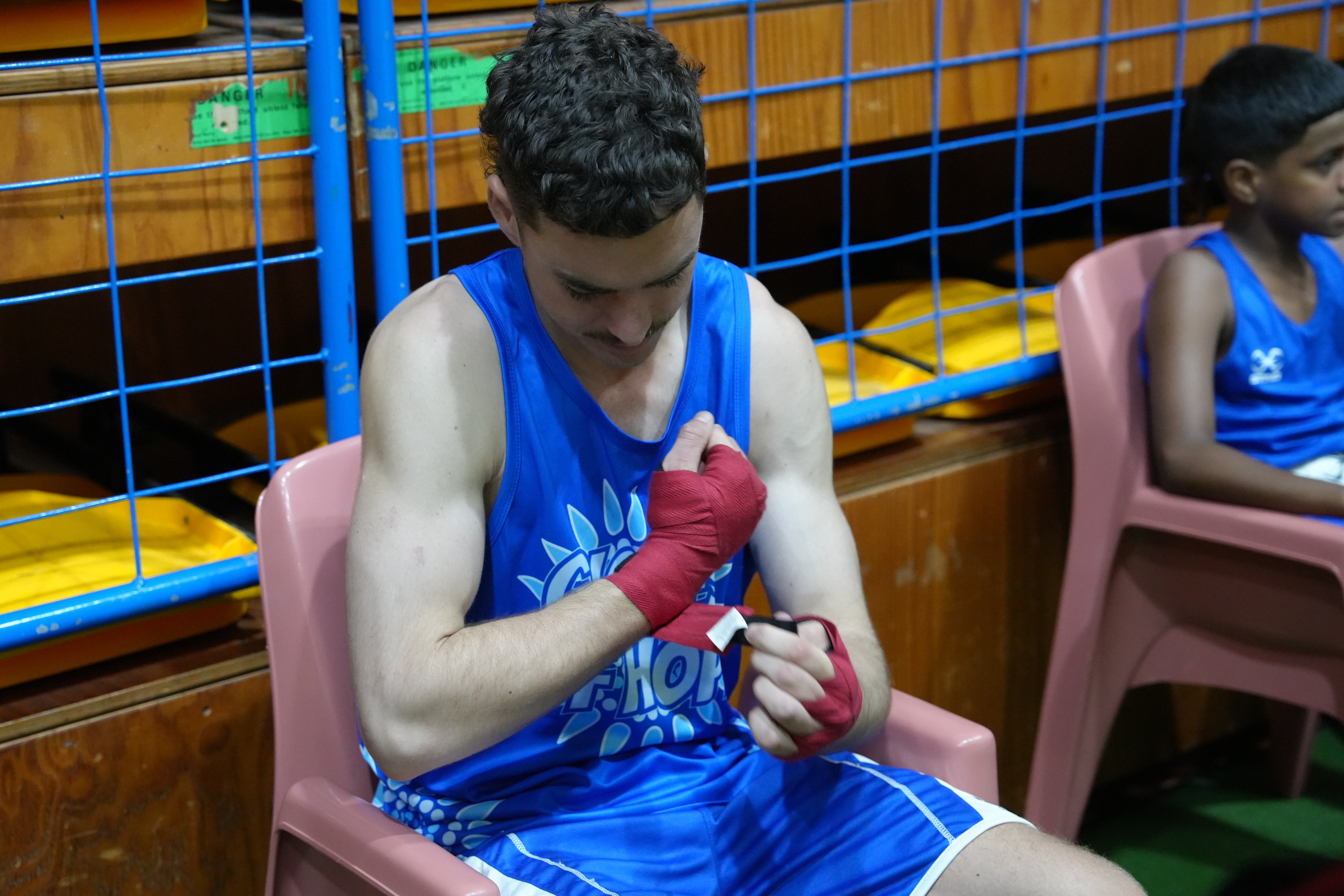 Declan Unwin-Walker wears a blue Gloves of Hope uniform inside a stadium, he wraps a strip of cloth around his hands and wrist.