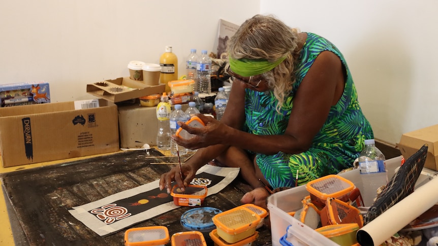 A woman sitting on the floor of a room and painting a canvas in a traditional Aboriginal style. 