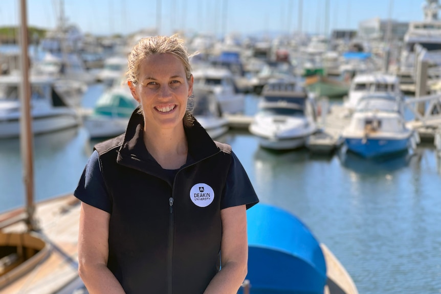 Profile picture of Dr Prue Francis, standing on a marina with a marina full of boats behind her.