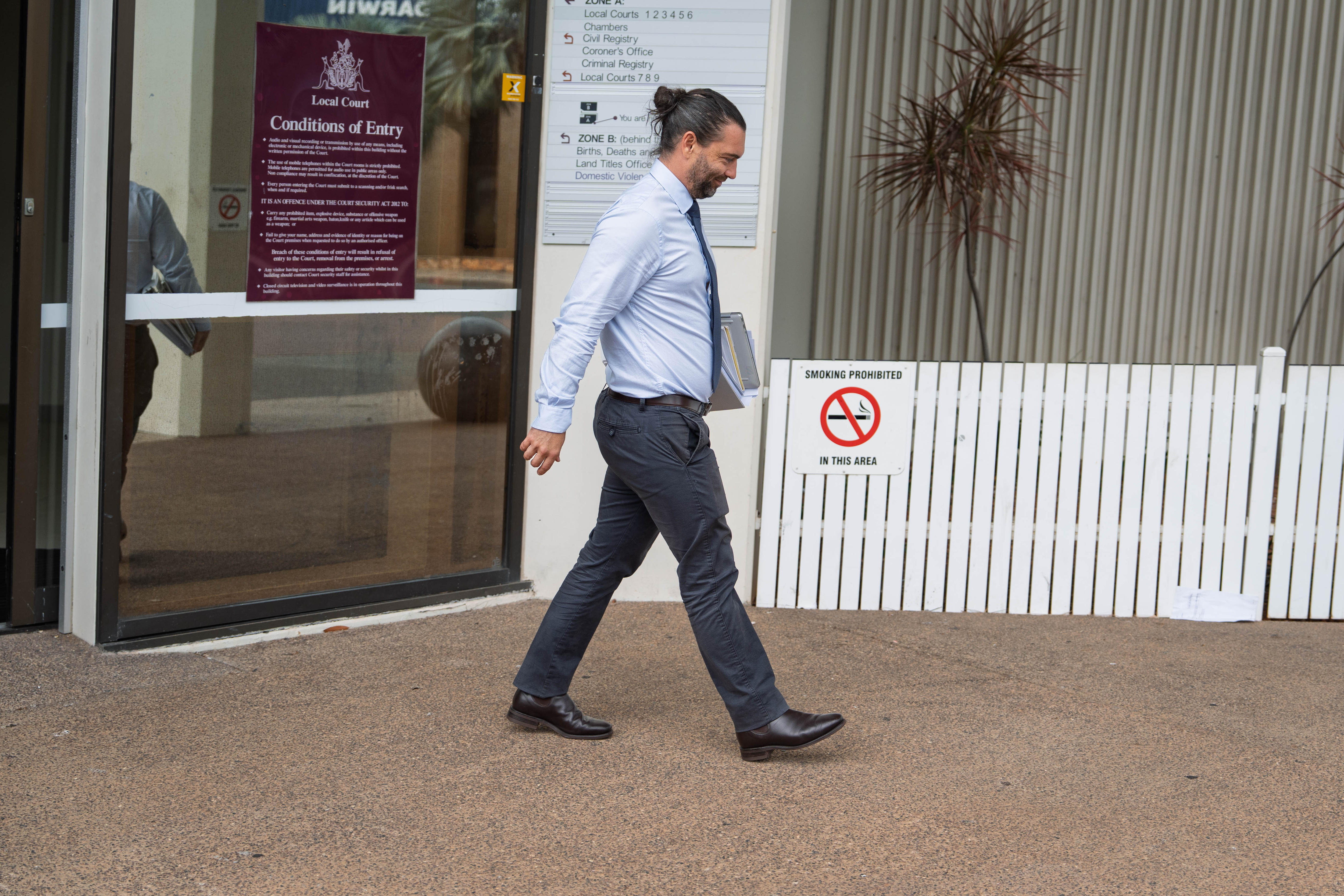 Um homem saindo da quadra, vestido com camisa azul, gravata e calça cinza. Ela tem um penteado com coque preto e está segurando documentos.