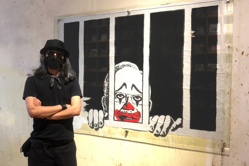A Malaysian man with long dark hair dressed in all black with a face mask, stands beside street art of Najib Razak as a clown.