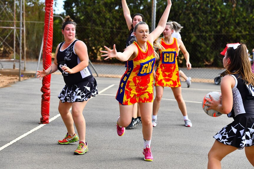 Netball players in action on a court wearing navy and white and red and yellow outfits.