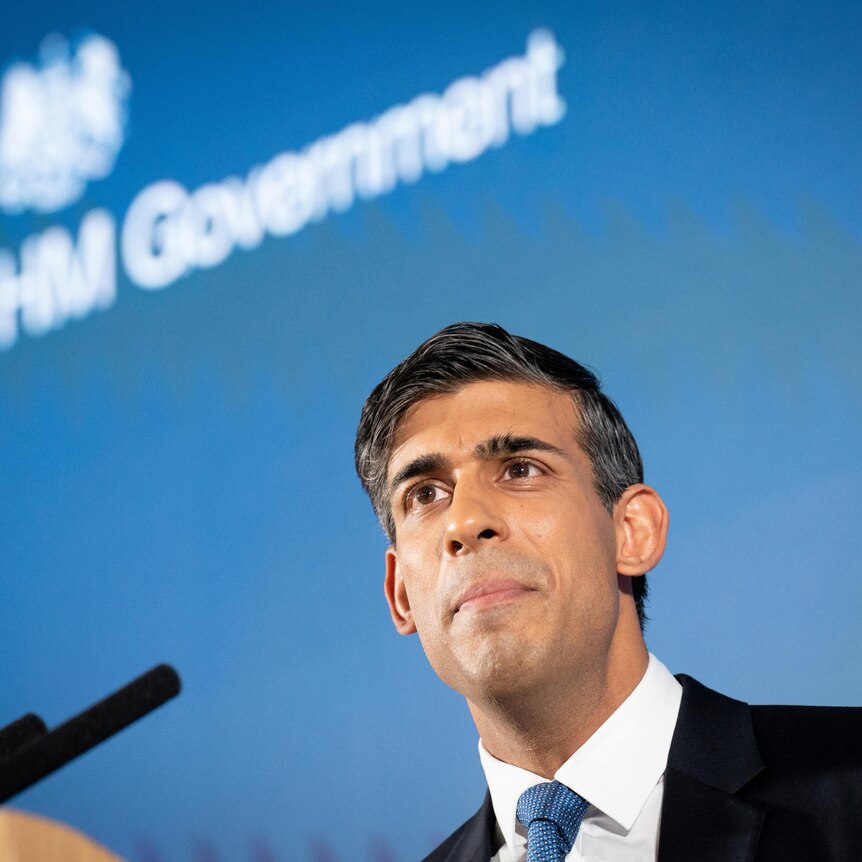 Sunak stands under text on a blue wall that reads "H M Government'
