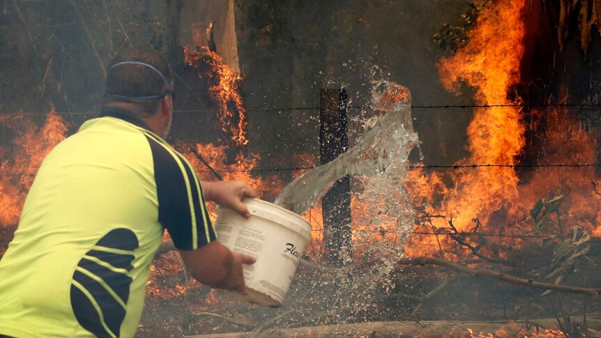 A man  in bright yellow t-shirt throws water on a bushfire with a bucket.