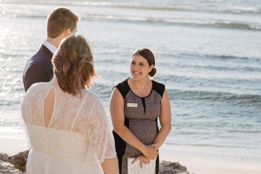 A woman with a bridal couple on a beach, bride viewed from behind.