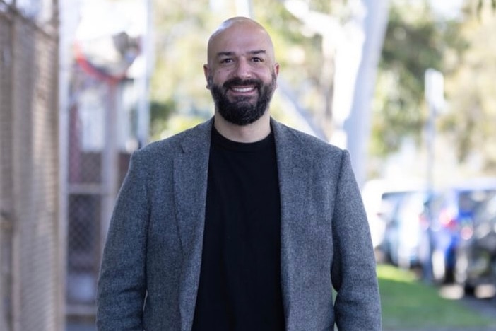 A photo of Andrew, wearing a grey jacket and black shirt, smiling, with a leafy street and cars visible in the background.