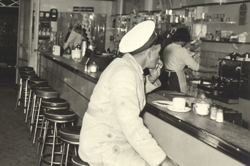 A cafe from 1952, with a milkman sitting at the bench drinking coffee.